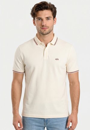 Hombre con cabello corto y castaño, vestido con una camiseta tipo polo color crema con rayas granate en el cuello y las mangas, de pie frente a un fondo liso.