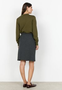 Olivengrön långärmad blus tillsammans med en knälång grå kjol med ett texturerat mönster och mörka blanka skor.
