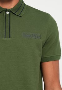 Grønn polo-shirt laget av strukturt stoff, med krage med marineblå striper, en mørkeblå brodert logo, og en to-knapp skjortepike.