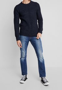 Laivastonsininen neulepaita, jossa on joustinneuletakki, pyöreä kaula-aukko ja pitkät hihat, yhdistetty kuluneisiin sinisiin farkkuihin ja mustiin lenkkareihin.