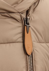 Detail z béžové prošívané látky s kovovým zipem a hnědým koženým táhlem s vyraženým písmenem "F."