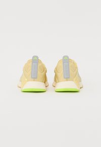 Scarpe da allenamento beige con tomaia in mesh, dettagli grigi e suola verde. Presentano un pannello di trazione sul tallone e elementi di design testurizzati.