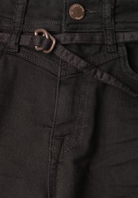 Close-up of black denim jeans with a thin fabric belt threaded through the loops and a metal button engraved with brand text.