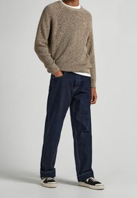 Maglione lavorato a maglia marrone con collo rotondo, abbinato a jeans larghi di colore blu scuro e sneakers nere con suola bianca.