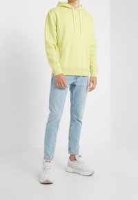 Světlá zelená mikina s kapsou na přední straně a šňůrkou na kapuci, v kombinaci s světlými modrými slim-fit džíny a bílými teniskami s akcenty.