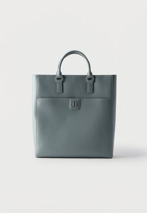 Sac cabas en cuir gris avec deux anses et une poche avant ornée d'un petit emblème logo métallique, posé sur un fond blanc uni.