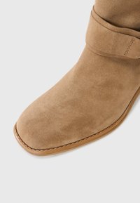 Botte cheville en suède beige avec un bout arrondi et une sangle ajustable autour du haut, sur une semelle marron contrastante.