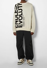 Pull en tricot beige avec texte noir "ENDLESS EVOLUTION." Associé à un pantalon noir ample et des baskets beige/noires.