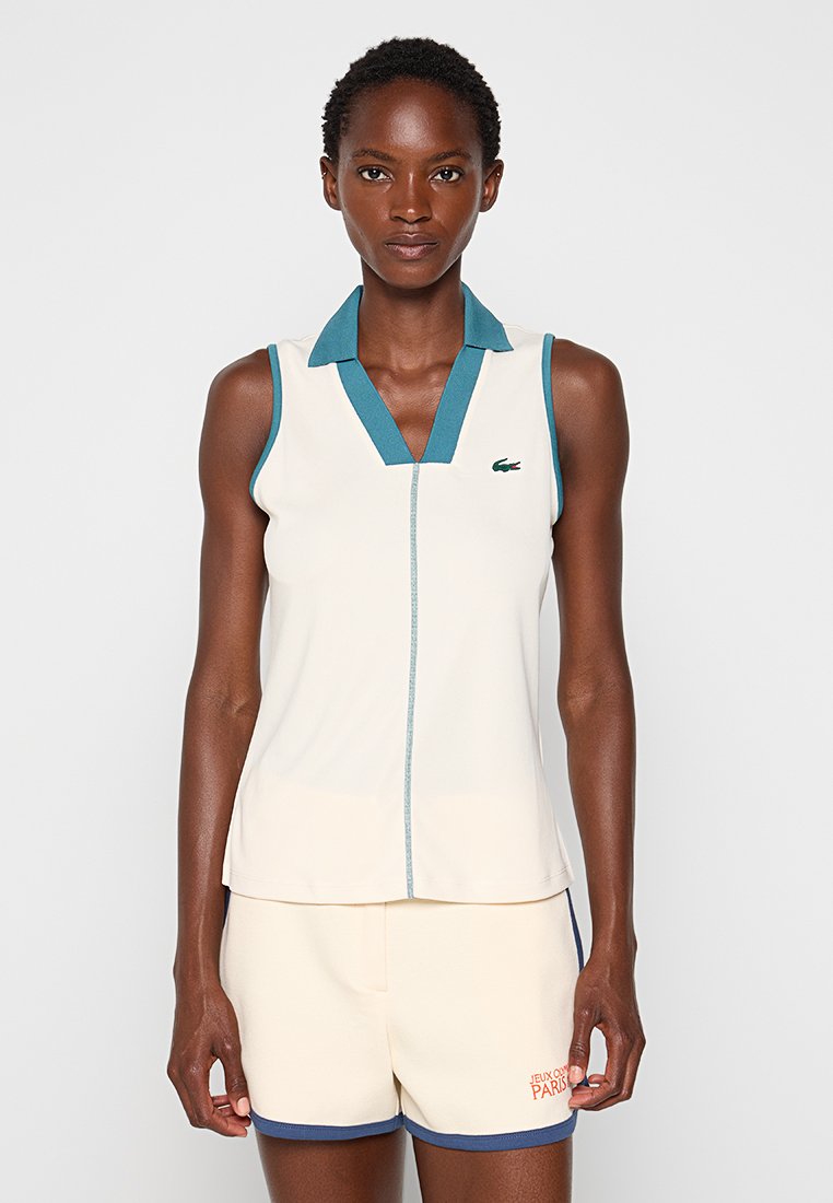 Ärmelloses cremefarbenes Polo-Top mit einem türkisfarbenen Kragen, verfügt über einen Reißverschluss vorne und ein grünes Logo auf der Brust. Glatte Textur, athletische Passform.