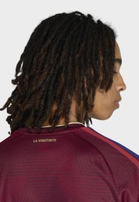 Maroon soccer jersey with geometric pattern; gold text "LA VINOTINTO" at the back; contrasting accents on shoulders. Textured fabric.