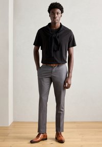 Camisa negra de manga corta con un suéter de punto oscuro anudado al cuello, pantalones gris ajustados y zapatos de vestir de cuero marrón, de pie sobre un suelo de madera.