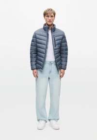 Kék puffertakaró dzseki vízszintes steppelt részekkel, magas gallérral és cipzáras zsebbel. Világoskék farmernadrággal és fehér tornacipővel párosítva.