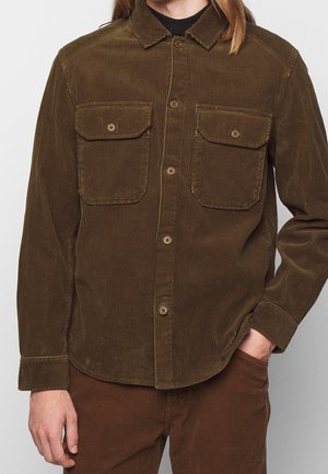 Homme portant une chemise en velours côtelé marron boutonnée avec deux poches à rabat à l'avant et un pantalon en velours côtelé marron assorti, main dans la poche.