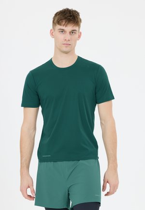 Ung mand iført en mørkegrøn sports-T-shirt med korte ærmer og matchende shorts, der står mod en ensfarvet hvid baggrund.