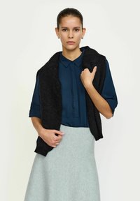 Zwarte, gebreide mouwloze vest over een blauwe blouse met knoopdetails, gecombineerd met een lichtgrijze, gestructureerde rok. Eenvoudige, elegante outfit.