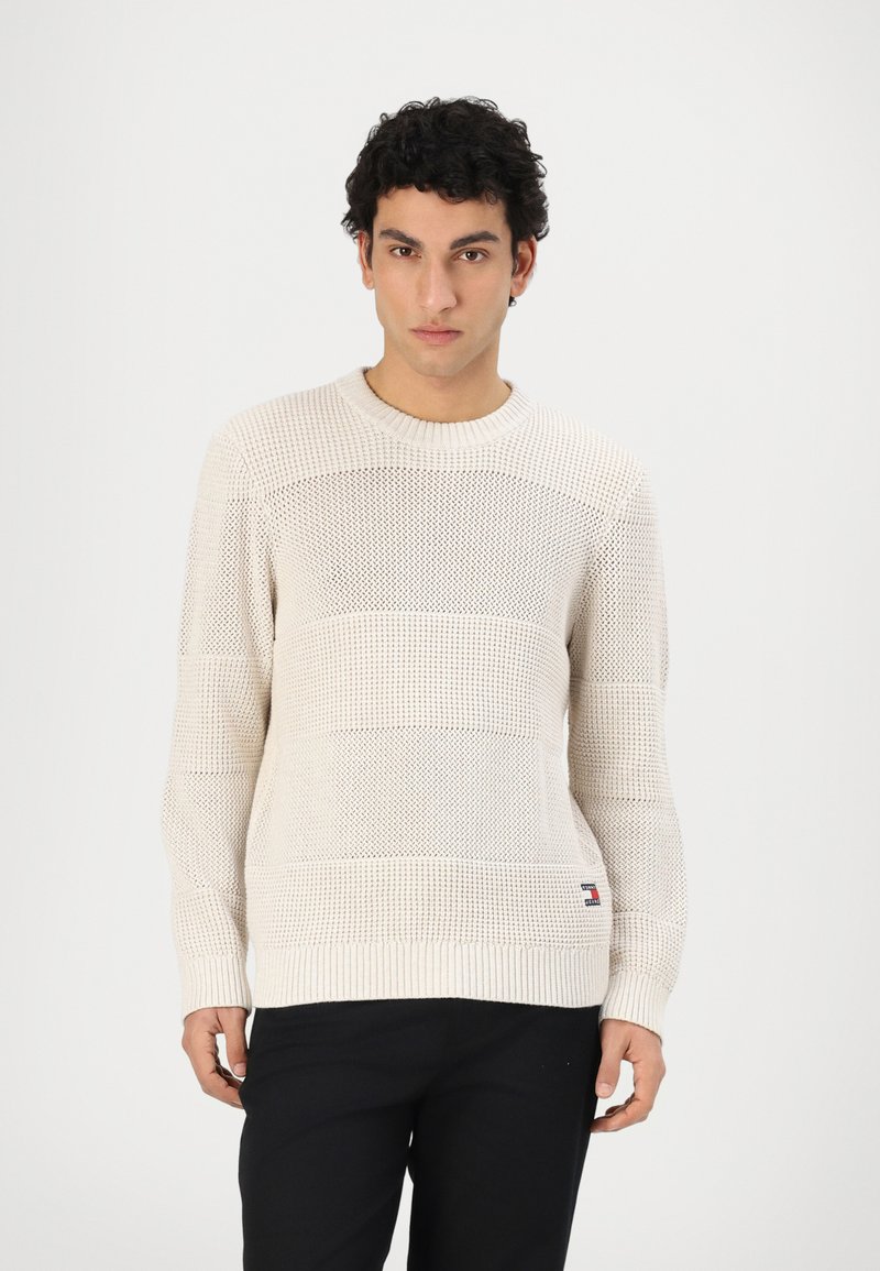 Suéter de punto en color crema con un diseño texturizado y estampado. Presenta un cuello redondo y puños de canalé, combinado con pantalones oscuros.