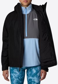 Czarna ocieplana kurtka z kapturem, noszona na jasnoniebieską i szaro-zieloną polarową bluzę z zamkiem. Model nosi niebieskie leggings w marmurkowy wzór.