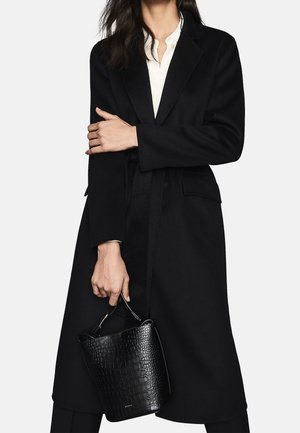 Femme portant un long manteau noir par-dessus une chemise blanche, tenant un sac à main noir texturé par sa poignée sur un fond uni.
