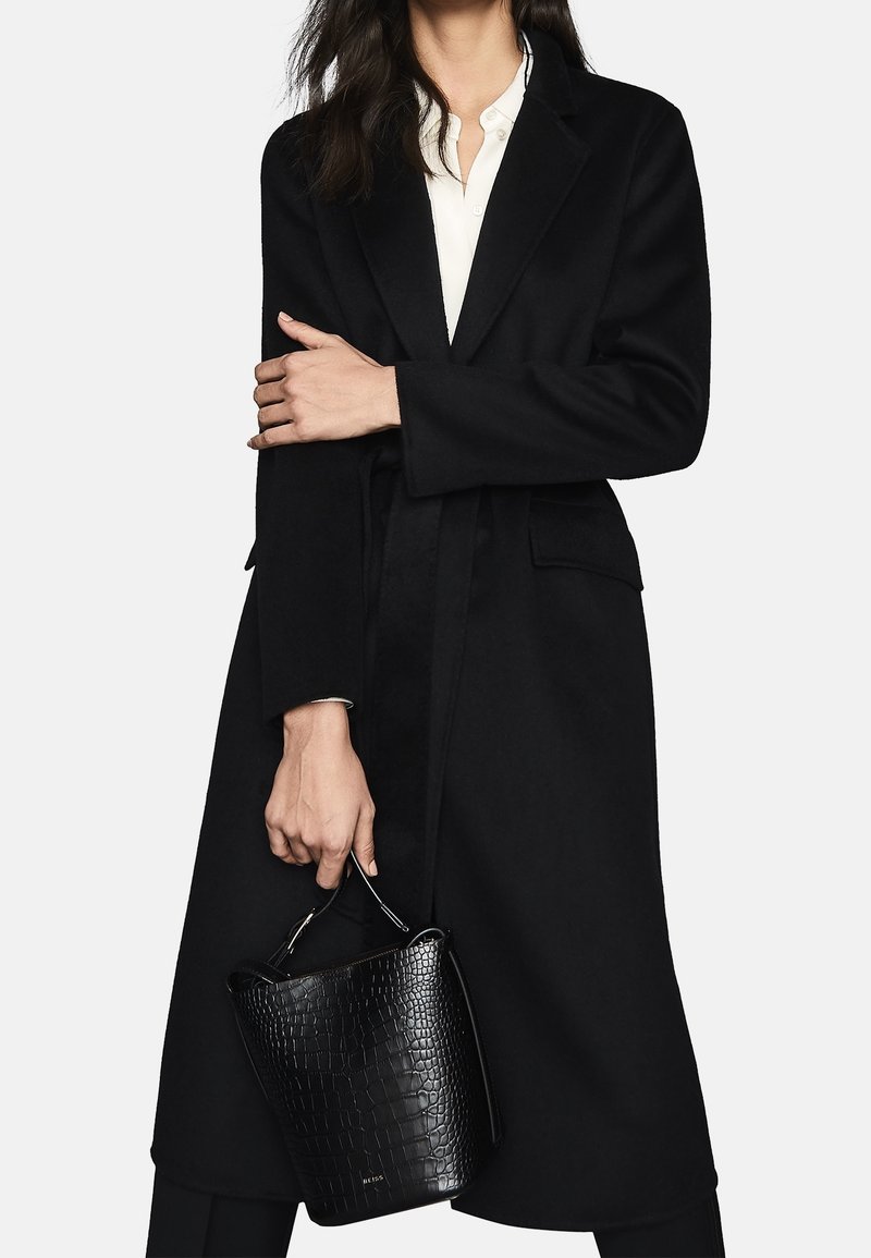 Femme portant un long manteau noir par-dessus une chemise blanche, tenant un sac à main noir texturé par sa poignée sur un fond uni.