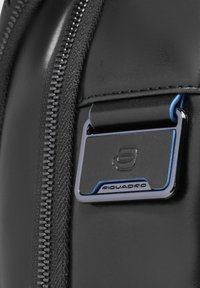 Zaino in pelle nera con una cerniera argento, caratterizzato da una targhetta metallica con marchio evidenziata in blu e una texture liscia.