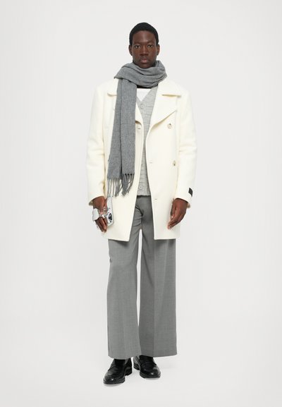 Cappotto bianco doppiopetto, sciarpa grigia, maglione grigio a trama, pantaloni ampi grigi, scarpe nere, in mano una piccola borsa argentata. Sfondo semplice.