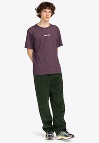 Camiseta de algodón morada con cuello redondo y logo "element" blanco, combinada con pantalones de pana verdes y zapatillas multicolor.