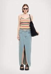 Gestreiftes ärmelloses Oberteil in Gelb, Rot und Schwarz, kombiniert mit einem hellblauen Jeans-Maxi-Rock mit einem Schlitz vorne. Schwarze Plateau-Sandalen.