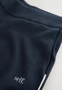 Pantalon de survêtement bleu marine avec une ceinture côtelée et un petit logo texte blanc portant l'inscription "self" sur le bas gauche. Texture de tissu douce.