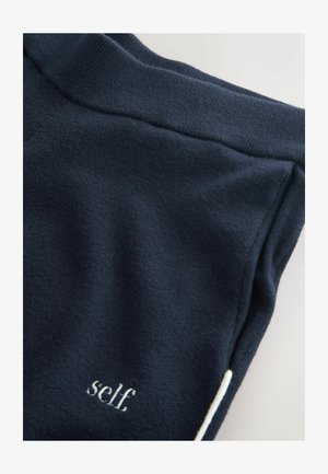 Pantaloni de trening bleumarin cu talie elastică și un mic logo text alb cuvântul „self” pe partea stângă jos. Textură moale a materialului.