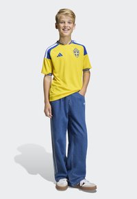 Gul kortärmad tröja med blå accenter och Sveriges nationella emblem. Kombinerad med löst sittande blå jeans och beige sneakers.
