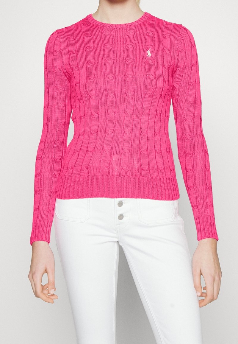 Suéter de punto rosa con patrón de trenzas, cuello redondo y puños y dobladillo acanalados. Pequeño logo en el lado izquierdo del pecho. Combinado con jeans blancos.