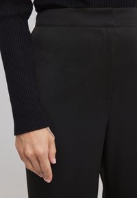 Hand med ljusrosa naglar och små tatueringar som vilar bredvid svarta högmidjade byxor och en svart ribbad långärmad topp.