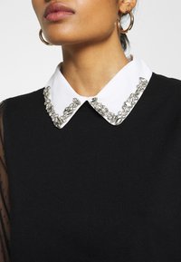 Svart tröja med en vit krage prydd med klara dekorationer och ett genomskinligt, prickigt tyg på ärmarna.