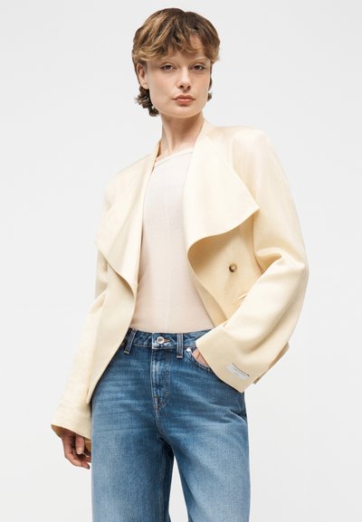Νεαρό άτομο με κοντά μαλλιά, φορώντας ένα μπεζ σακάκι με draped λεπτομέρειες πάνω από ένα μπεζ τοπ και μπλε τζιν, στέκεται με το ένα χέρι στην τσέπη.