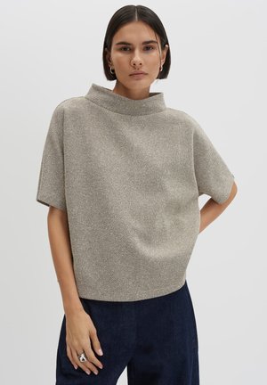 Vrouw met kort donker haar, die een gestructureerde beige korte mouwen top en een donkere broek draagt, staand tegen een effen lichte achtergrond.