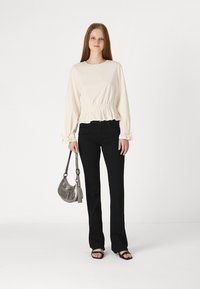 Jeune femme portant un haut blanc à manches longues avec une taille froncée, un pantalon noir, des sandales noires, tenant un petit sac à main en argent, se tenant sur un fond blanc.