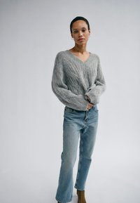 Frau steht vor einem schlichten Hintergrund und trägt einen grauen V-Ausschnitt-Pullover mit Zopfmuster, hellblaue Jeans und braune Knöchelstiefel.