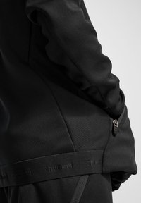 Svart idrottsjacka med texturerad tyg, med en dragkedja sidoficka och ett märkes elastiskt band vid fållen. Slät finish.