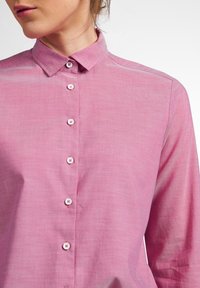 Žena v růžové košili s dlouhým rukávem a límečkem, s nadepnutými knoflíky, na bílém pozadí.