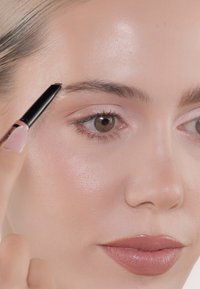 Close-up van een persoon die een zwarte wenkbrauwpotlood aanbrengt. Heeft een egale huid, lichte blush en neutrale lippenstift op een lichte teint.