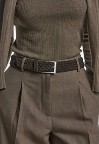 Close-up van een persoon in een bruine geribbelde trui die is ingestopt in bruine plooibroeken met een donkere gevlochten riem om de taille.