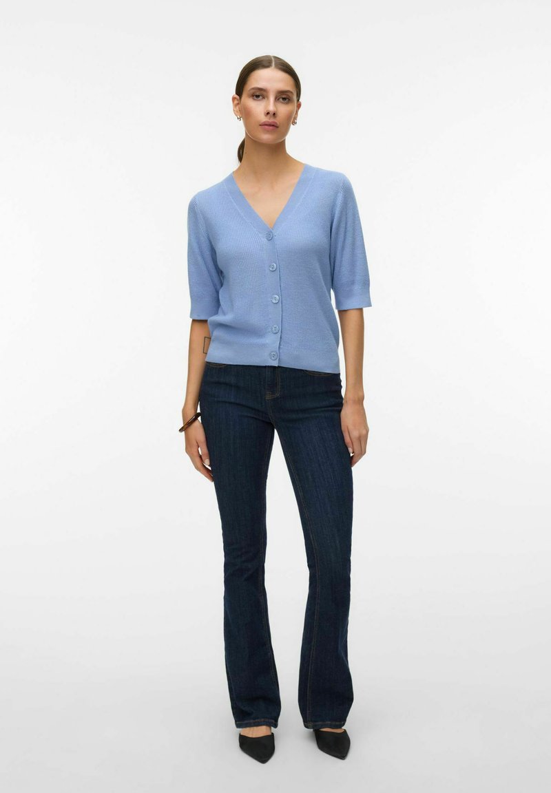 Femme portant un cardigan boutonné à manches courtes bleu clair associé à un jean évasé foncé et des chaussures noires à bouts pointus, debout devant un fond blanc.
