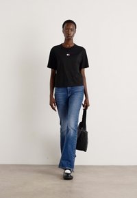 T-shirt negru cu mâneci scurte și un logo mic, asociat cu blugi evazați de culoare albastru deschis și pantofi negri. O geantă de umăr este ținută într-o mână.