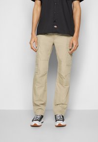 Beige Cargo-Hosen mit mehreren Taschen, aus strapazierfähigem Stoff. Getragen mit einem schwarzen Hemd und schwarzen Sneakers mit weißen Akzenten.