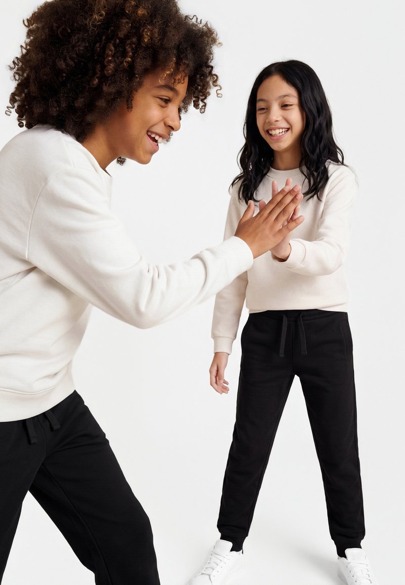 To smilende børn iført hvide toppe og sorte bukser giver hinanden et high five mod en ensfarvet hvid baggrund.