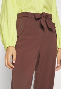 Kvinna i bruna högmidjade byxor med knuten bälte och fickor med klaff, kombinerat med en ljusgrön långärmad blus med samlade ärmslut.