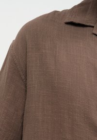 Καφέ υφασμάτινο λινό πουκάμισο με υφή, με ορατό ραφή στον ώμο και μερικό γιακά πάνω σε ουδέτερο φόντο.