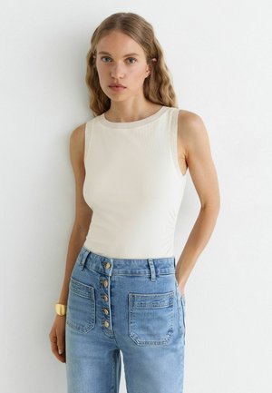 Jonge vrouw met lang blond haar die een mouwloze witte top en een high-waist blauwe spijkerbroek met knopen aan de voorkant draagt, staand tegen een witte muur.