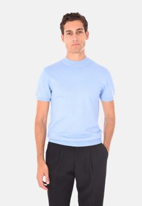 Uomo con capelli ricci corti che indossa una maglietta a maniche corte di maglia azzurra e pantaloni neri plissettati, in piedi davanti a uno sfondo bianco.