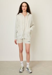 Jasnoszara bluza rozpinana z kapturem i sznureczkami, zestawiona z dopasowanymi szortami z naszywką z logo. Model nosi skarpetki w wzory i buty do biegania.